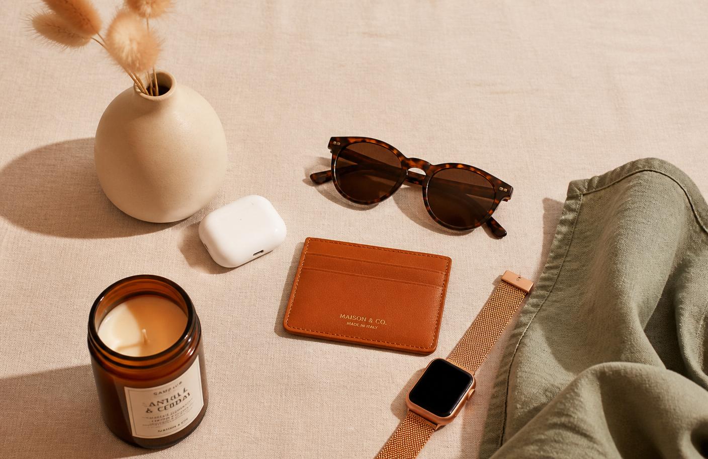 A curated flatlay of trending lifestyle products including sunglasses, a smartwatch, earbuds, a leather wallet, a candle, and a ceramic vase